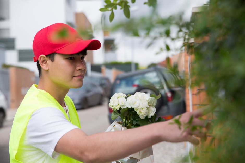 A young delivery person wearing a red cap and a high-visibility yellow vest is seen arranging a fresh bouquet of white hydrangeas and roses against a brick wall with a background of parked cars and re