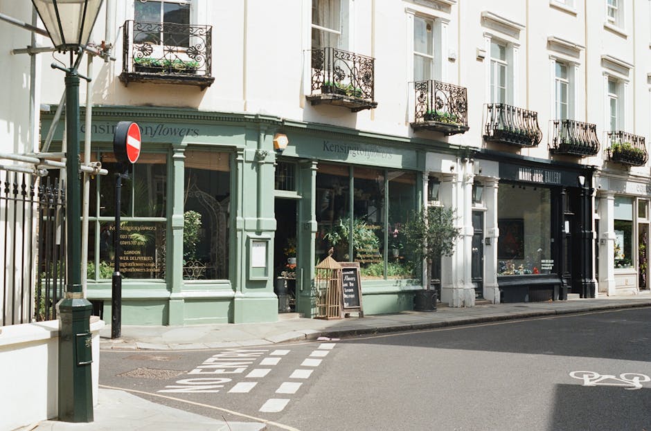 A row of quaint floral shops with large display windows showcasing fresh flowers in various colours, including pinks, reds, and whites, arranged in traditional bouquets and vases. The buildings are pa