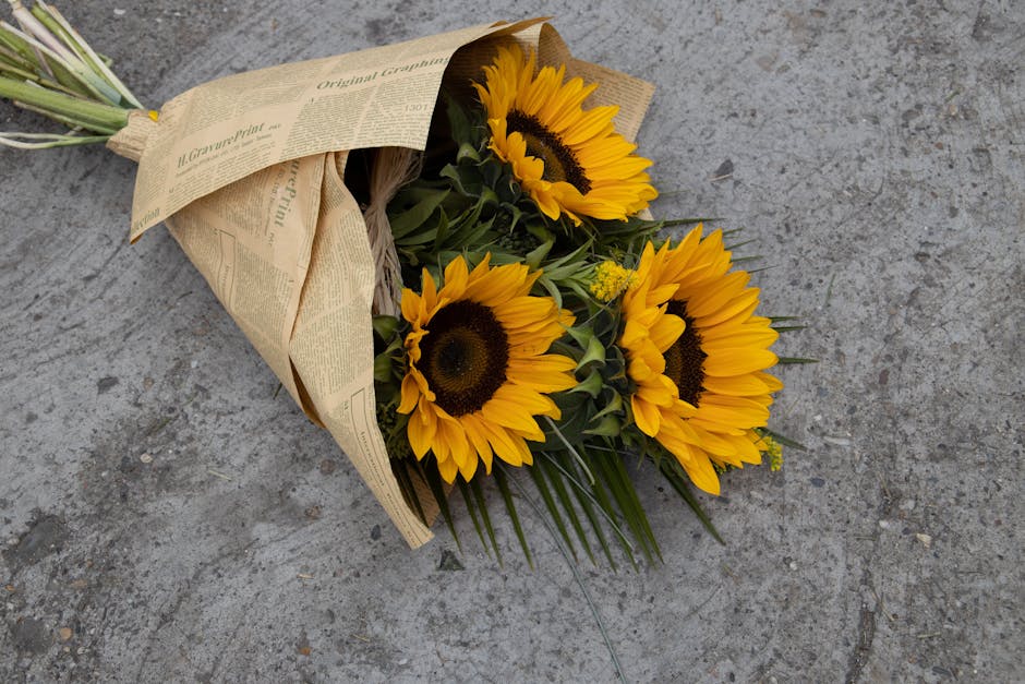 A bouquet of fresh, vibrant yellow sunflowers with dark centers, wrapped in rustic brown paper with a layer of newspaper print, resting on a gray concrete surface. The arrangement features green folia