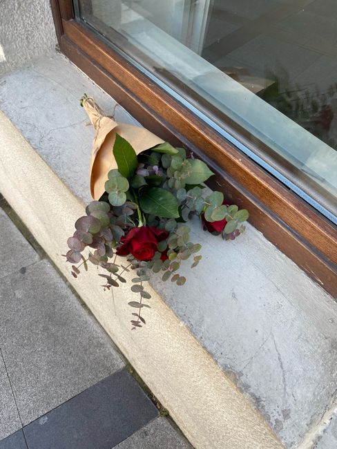 A single-stem red rose with deep red petals and a few green leaves, wrapped in light brown paper, rests on a concrete window ledge next to a large window with a wooden frame. Eucalyptus branches with 
