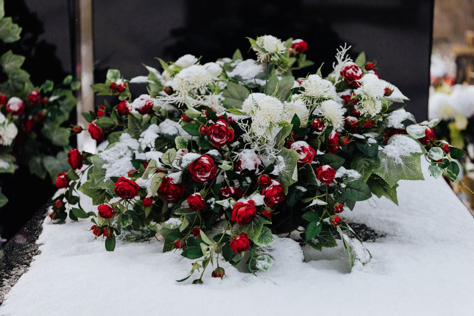 A floral arrangement of fresh red roses and white chrysanthemums, mixed with green holly leaves, placed on a snow-covered surface. The bouquet is partially dusted with snow, highlighting the vibrant c