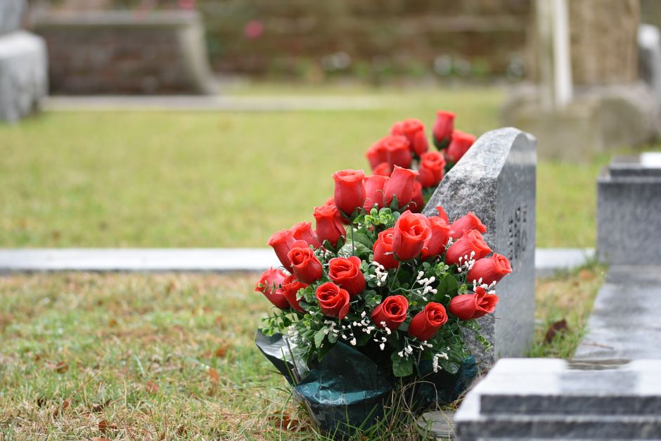 A bouquet arrangement of fresh red roses with green foliage and small white accent flowers, wrapped in dark green cellophane, rests against a grey granite gravestone in a cemetery. The bouquet is plac