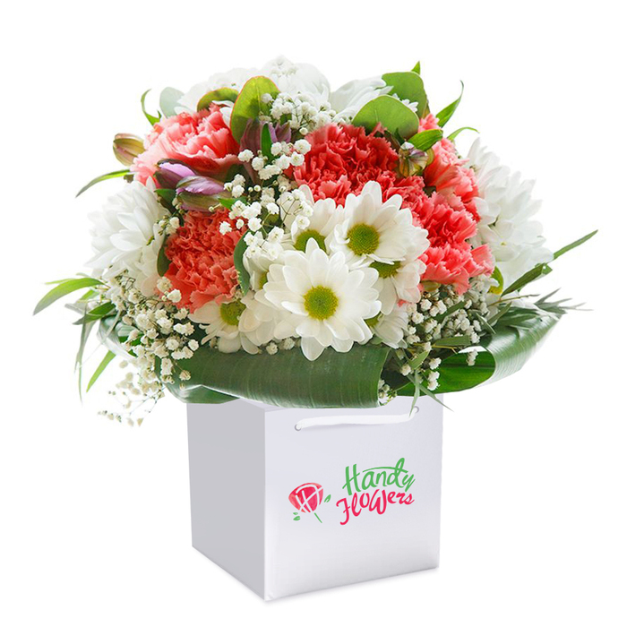 At the heart of this photograph is a striking hand-tied bouquet, poised in a modern white square gift bag, all set against a flawless studio white that allows the colours to sing. Each bloom within the arrangement has been chosen for harmony and contrast: coral and peach-toned carnations with gently ruffled petals unfurl beside clusters of crisp white daisies, their vivid chartreuse centers brightening the ensemble like the lively markets near Kensal Green station. Ethereal sprays of baby's breath are lightly scattered throughout, lending a delicate veil and letting each flower share the spotlight. Toward the left edge, tight buds tinged with purple suggest growth and continuity, adding subtle depth and movement. Encircling it all is a lush necklace of deep green leaves-broad, glossy, and meticulously placed for balance and flow, reminiscent of the leafy walks along Bosworth Road. The white gift bag, designed for both elegance and convenience, features durable white corded handles, perfect for gifting in NW10. Every detail, from the careful arrangement to the radiant finish of the petals, reflects the thoughtfulness and skill of a local florist who understands the character of Kensal Green. Ideal for marking life's joyful chapters or sending a thoughtful note, this bouquet's freshness and refined presentation convey a sense of hope and happiness unique to local London flower delivery.
